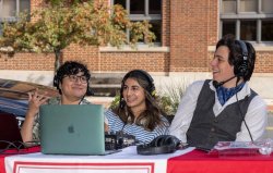 Three young students sitting outside at a table with a laptop in front of them. All of them are wearing headphones and are part of a live radio broadcast. They are smiling and happy.
